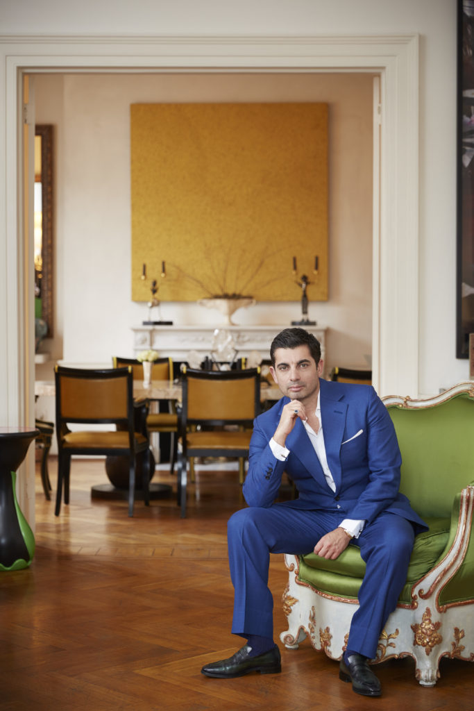 Designer Francis Sultana wears a blue suit and sits on a green, white and gold chair with a yellow painting in the background