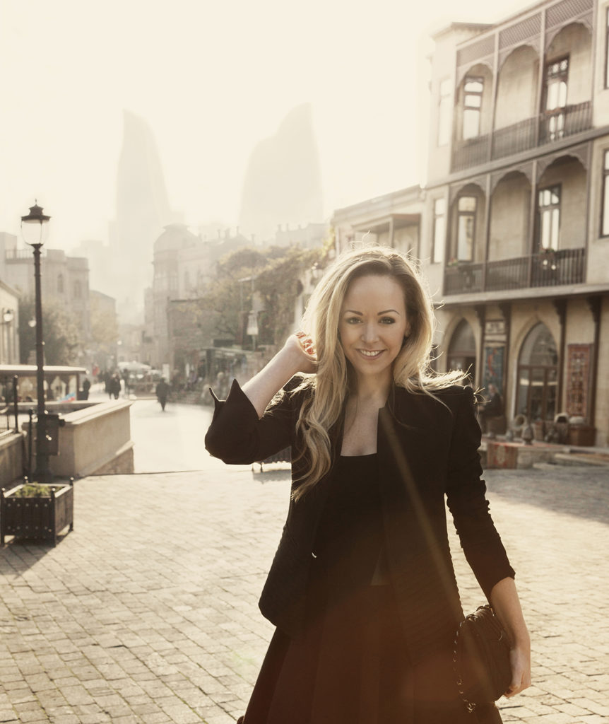 Tamara Ralph wears a black jacket and dress and bag in the Old Town in Baku
