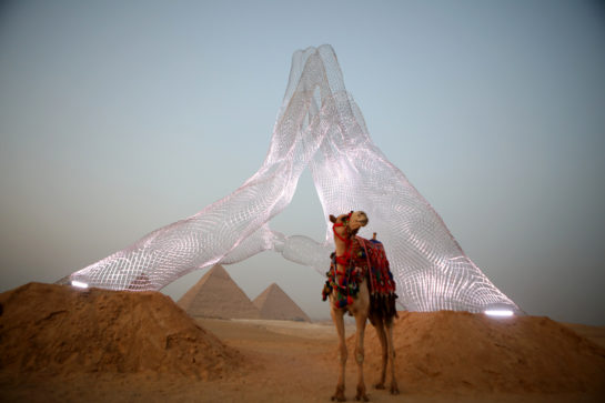 A camel in front of a sculpture and pyramids in a desert