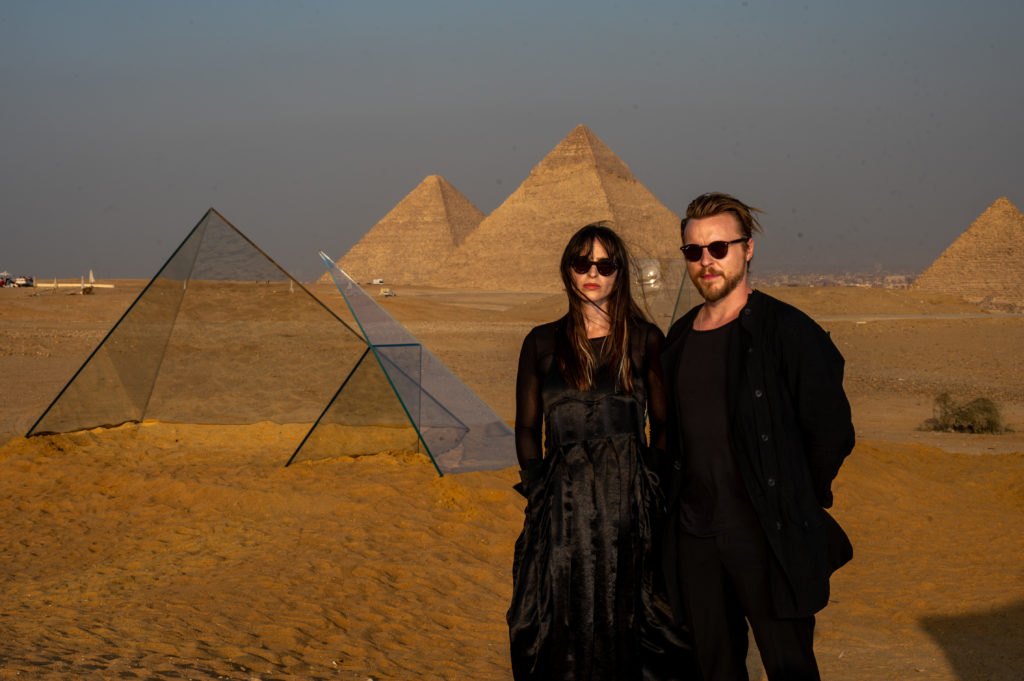 A man and woman both wearing black standing in front of mini glass pyramids next to real pyramids