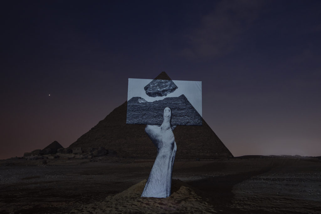A giant hand coming out of the ground holding a picture of a pyramid against a pyramid at night