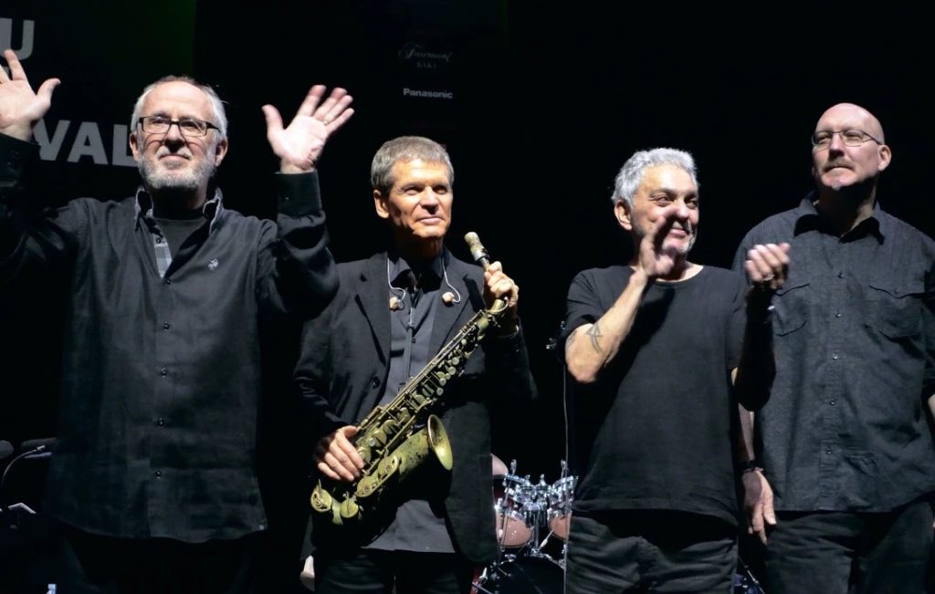 four men in black and one is holding a saxophone