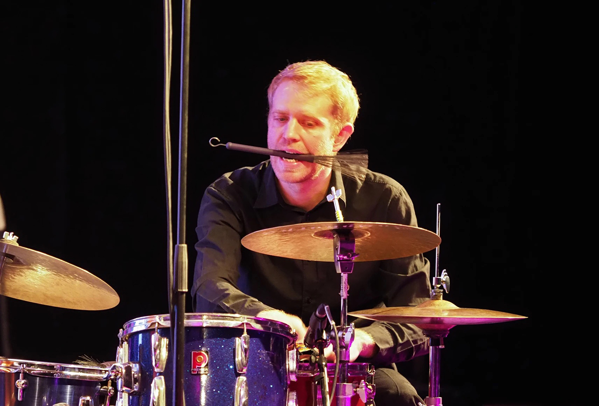A man playing on the drums with a shaker in his mouth