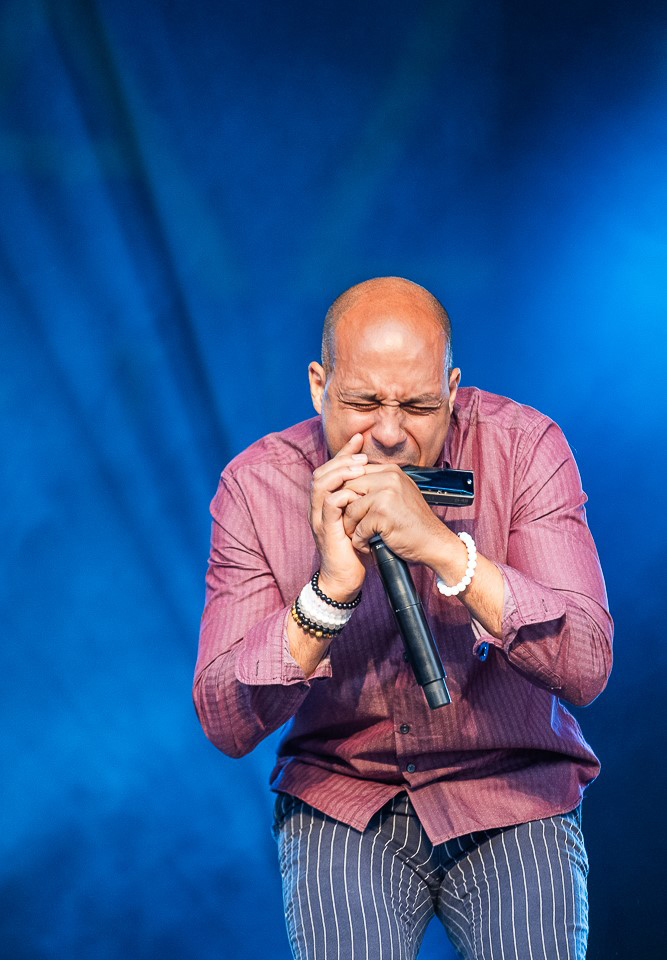 A man wearing a red shirt and blue and white striped trousers playing a harmonica passionately 