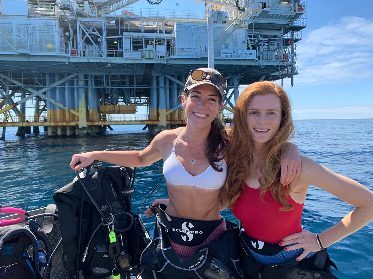 Two females on boat with scuba diving equipment, oil rig in background